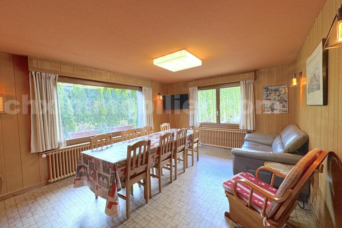 A cozy dining and living room featuring wood paneling, a long table with chairs, a sofa, and large windows overlooking green foliage.