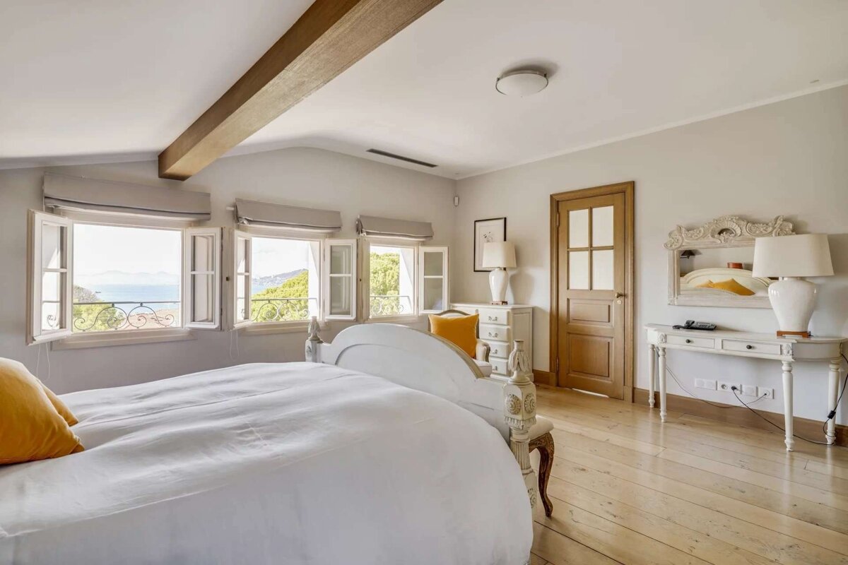 Bright bedroom with a white bed, light wood floor, and open windows revealing a scenic view of the sea and green landscape.