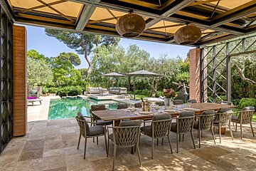 A beautiful covered outdoor dining area with a long table overlooking a sparkling swimming pool and lush green garden on a sunny day.