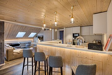 A cozy, modern open-plan kitchen and living space with warm wood ceilings & walls, an illuminated bar, stylish pendant lights, and skylights.