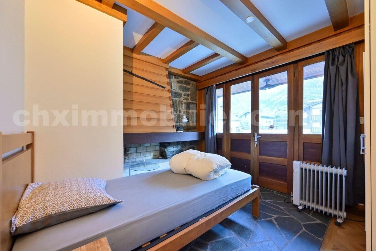 A rustic room features a single bed, wooden ceiling beams, a stone fireplace, and French doors with dark curtains. A stone floor and radiator are visible.