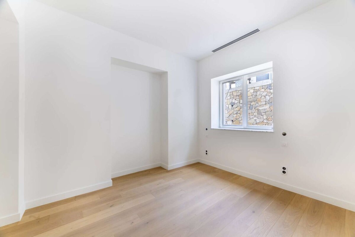 Bright, empty room with white walls, light wooden floor, a window revealing a stone wall, and a built-in alcove.
