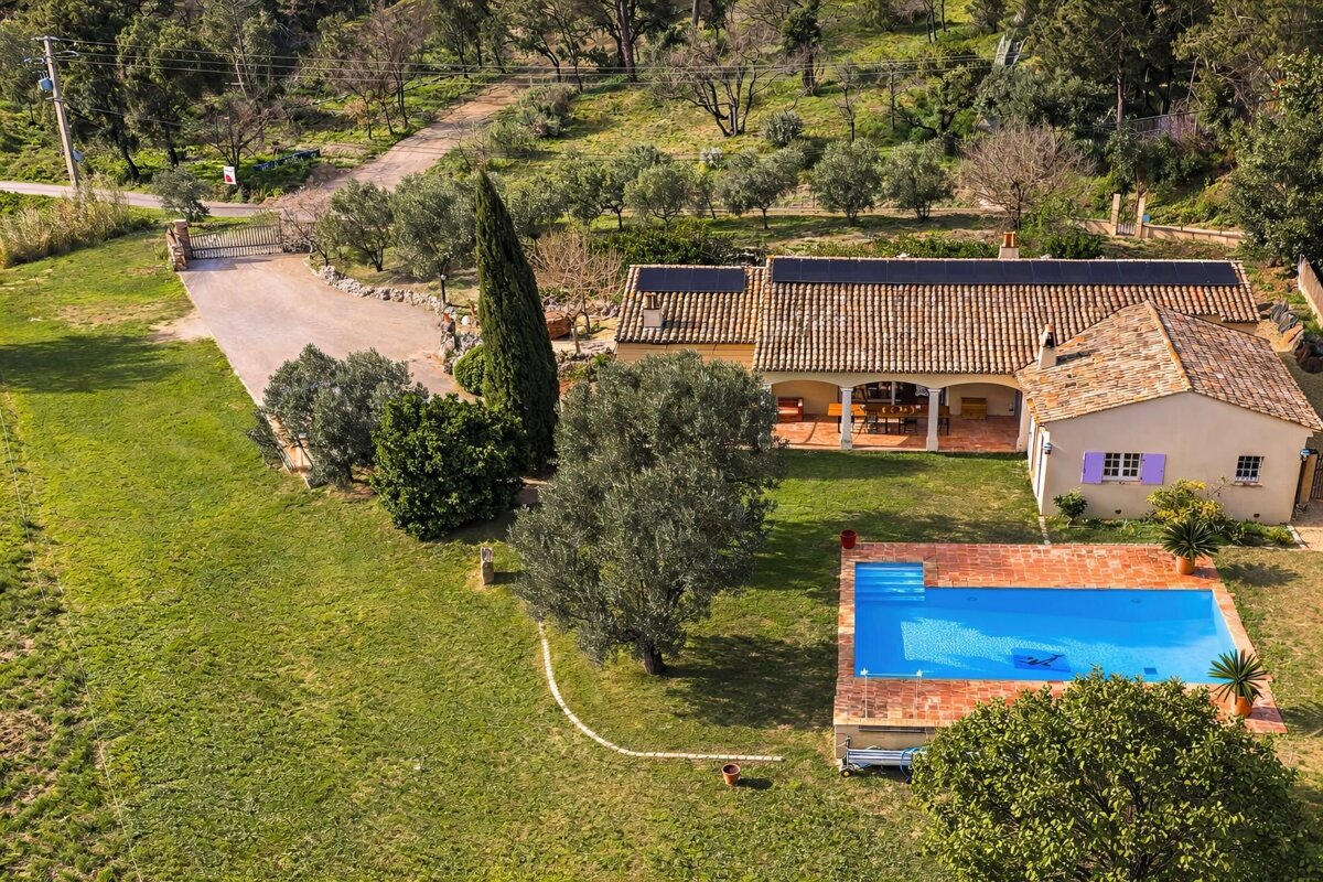 Aerial view of a charming villa with a tiled roof, sparkling blue swimming pool, lush green lawn, and olive trees. A driveway leads to the property.