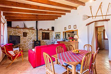 A rustic, cozy living/dining room features exposed wooden beams, a stone accent wall with a wood stove, and a vibrant red sofa. A wooden dining table with a striped tablecloth sits centrally.