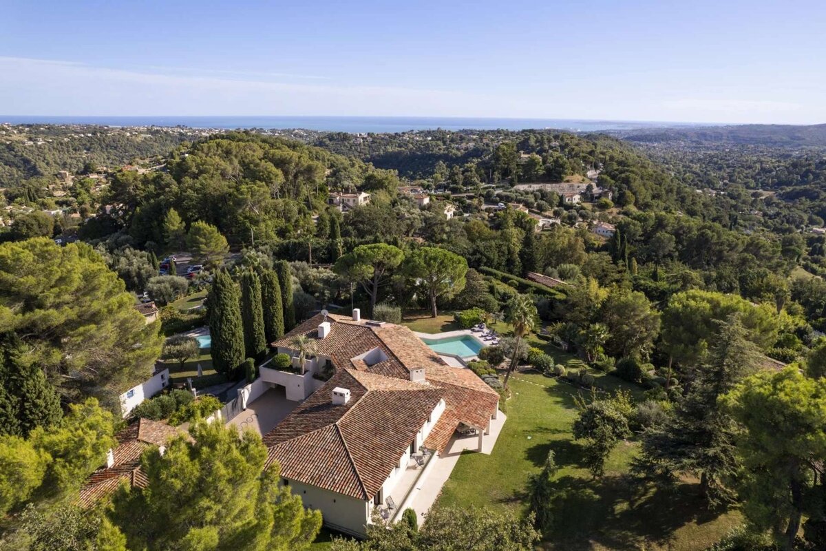 Aerial view of a large villa with a terracotta roof and swimming pool, nestled in a lush green landscape with hills and the sea in the distance.