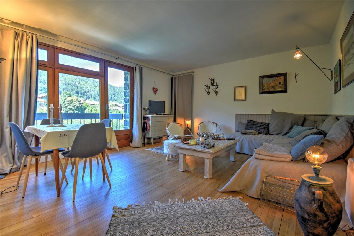 A rustic living room with a dining area, featuring wooden floors, a cozy sofa, and a large window overlooking a lush mountain landscape. Warm, inviting decor.