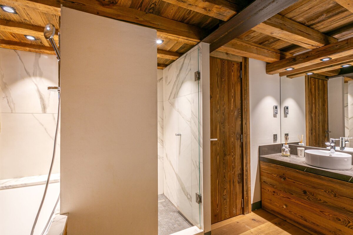 Stylish bathroom with a rustic wooden beamed ceiling, white marble shower, and a wooden vanity with a sink and large mirror.