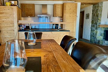A rustic wooden dining table with leather chairs, looking towards a warm kitchen with wooden cabinets in the background.