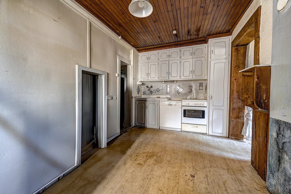 An empty, dated kitchen with white cabinets, appliances, a warm wooden ceiling, and a worn wooden floor. Plain white walls.