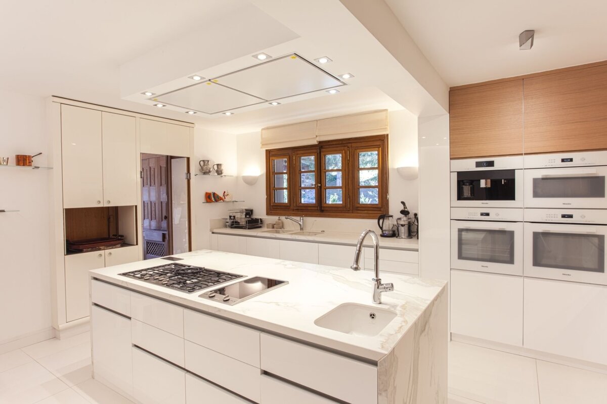A pristine, modern white kitchen featuring a large island with cooktop/sink, integrated ovens, and a prominent wood-framed window.