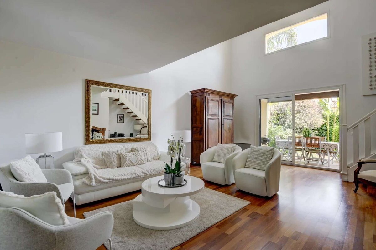 A bright living room features white sofas and chairs, a large mirror reflecting a staircase, and hardwood floors. A glass sliding door opens to a lush outdoor patio.