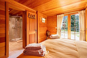 Cozy wooden chalet bedroom featuring a bed with folded towels and an open doorway leading to an en-suite bathroom with a glass shower.