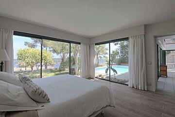 Modern bedroom with a white bed and floor-to-ceiling windows overlooking a swimming pool, lush garden, and the sea. Two crane statues are by the pool.