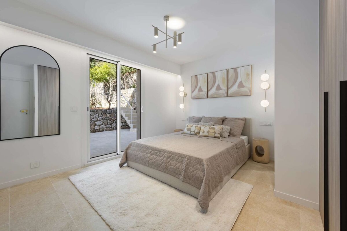 A bright, modern bedroom with white walls, neutral bed, arch mirror, contemporary lighting, and sliding glass doors opening to an outdoor patio.