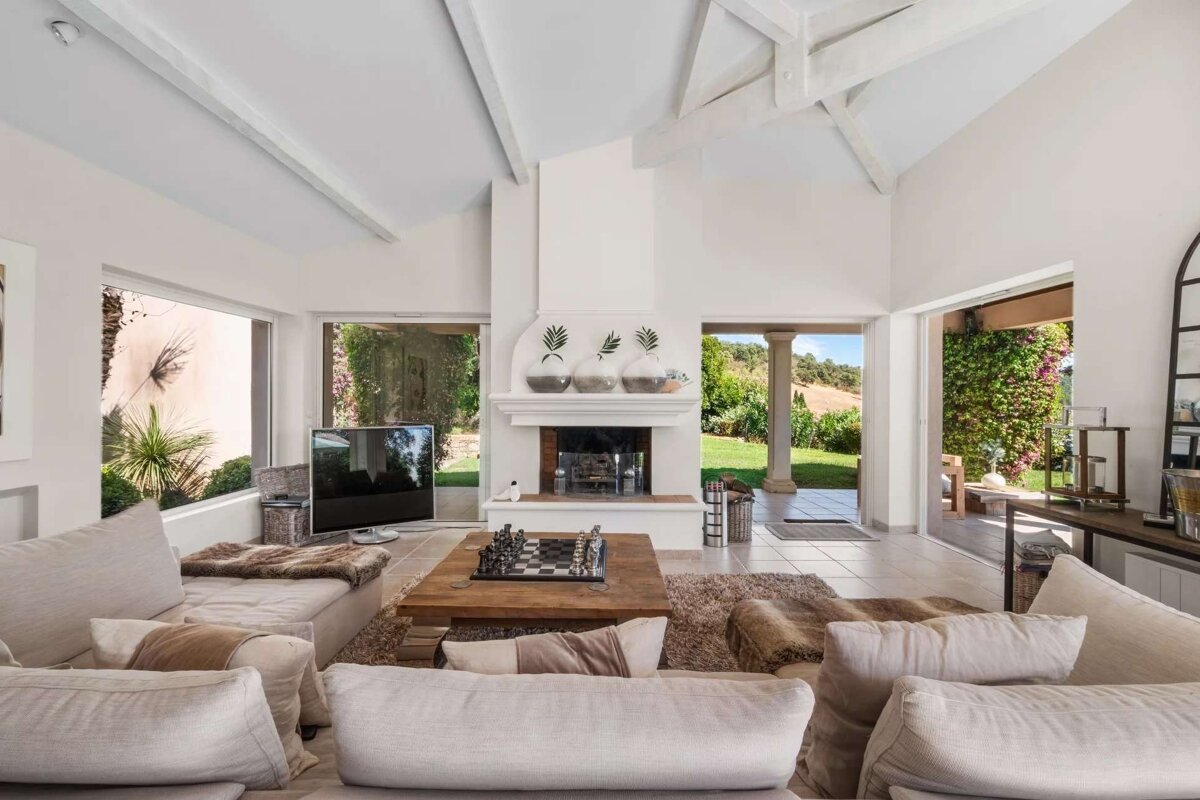 A bright, spacious living room with high beamed ceilings, plush light sofas, a fireplace, and a chess set on a coffee table, opening to a lush green garden.
