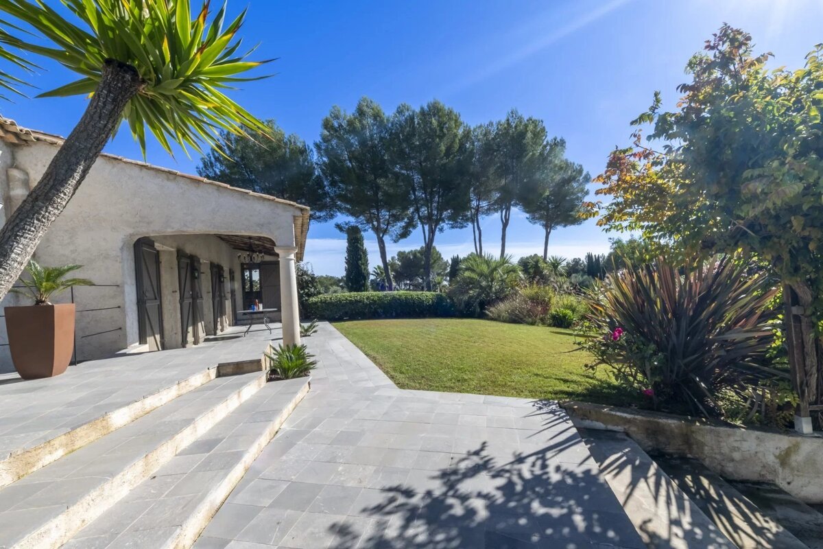 Sunny villa patio with stone steps, a lush green lawn, and tall trees under a clear blue sky.