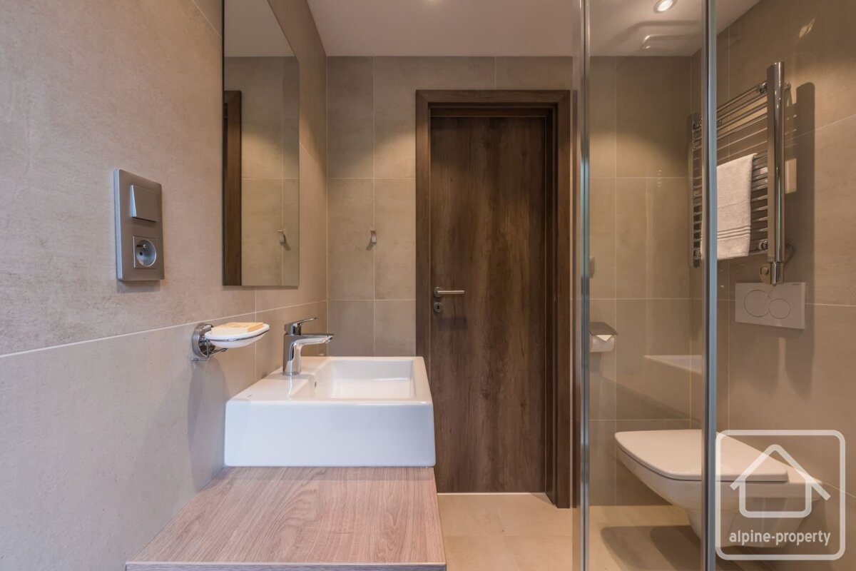 A modern bathroom featuring a white sink on a wooden counter, tiled walls, a wooden door, and a glass shower enclosure with a toilet and towel warmer.