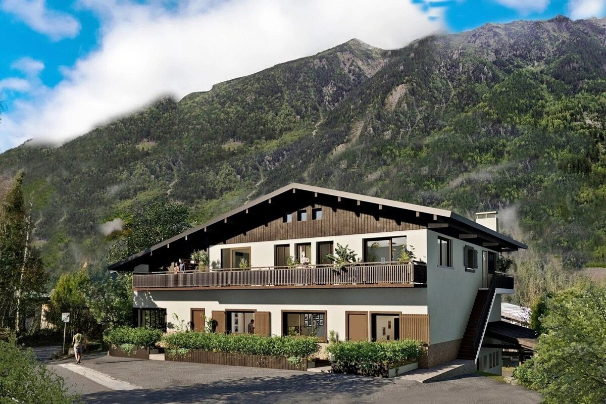 A two-story mountain chalet with a wide balcony, set against a massive, lush green mountain under a blue sky.