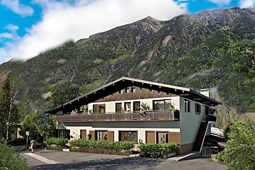 A two-story mountain chalet with a wide balcony, set against a massive, lush green mountain under a blue sky.