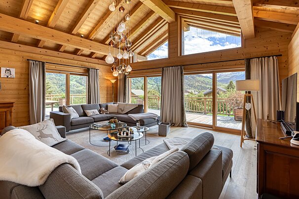 A cozy, modern chalet living room with wooden interior, large grey sofas, a unique chandelier, and vast windows overlooking a stunning mountain vista.