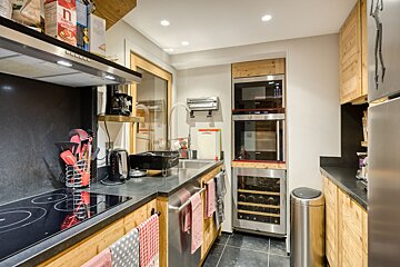 A modern kitchen with light wooden cabinets, dark countertops, and stainless steel appliances including an oven, microwave, wine cooler, and cooktop.