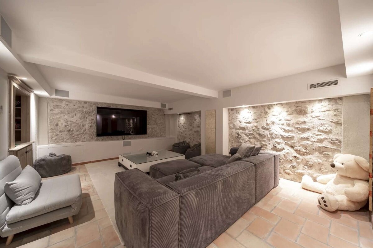 A cozy media room with exposed stone walls, a large TV, grey sectional sofas, terracotta tile floors, and a huge white teddy bear.