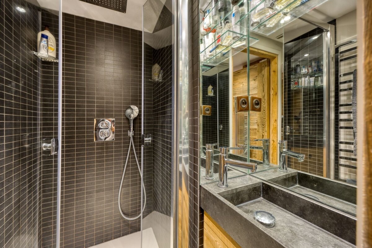 A sleek, modern bathroom features a dark tiled shower with glass door, alongside a stone double-sink vanity with mirrored cabinets reflecting wooden walls.