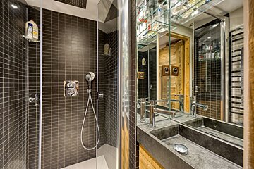 A sleek, modern bathroom features a dark tiled shower with glass door, alongside a stone double-sink vanity with mirrored cabinets reflecting wooden walls.