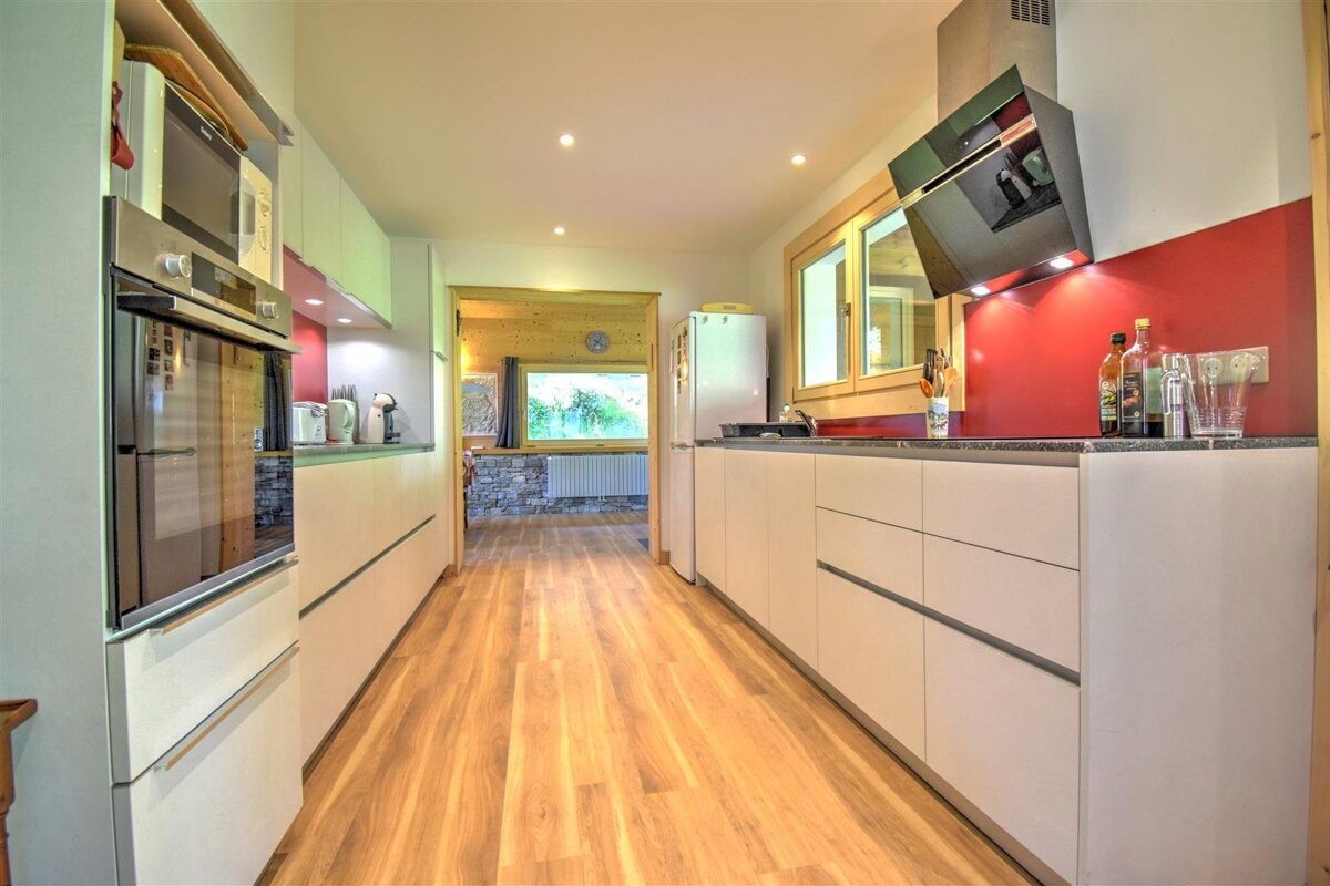 A modern galley kitchen featuring sleek white cabinets, a bold red backsplash, and warm wood flooring. It's well-lit with natural light and recessed spotlights.