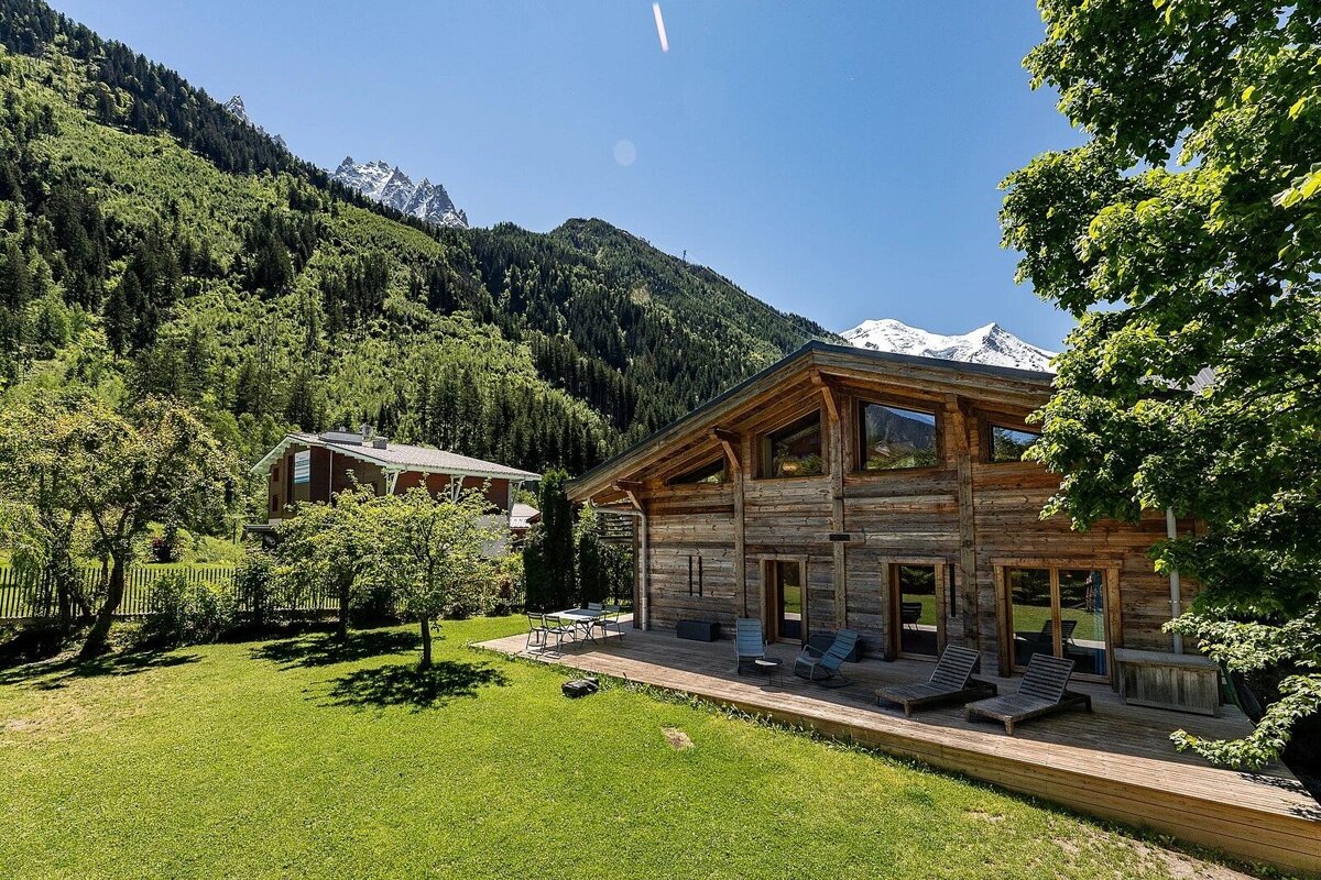 A rustic wooden chalet with a green lawn and deck, set against lush forested mountains and snow-capped peaks under a clear blue sky.