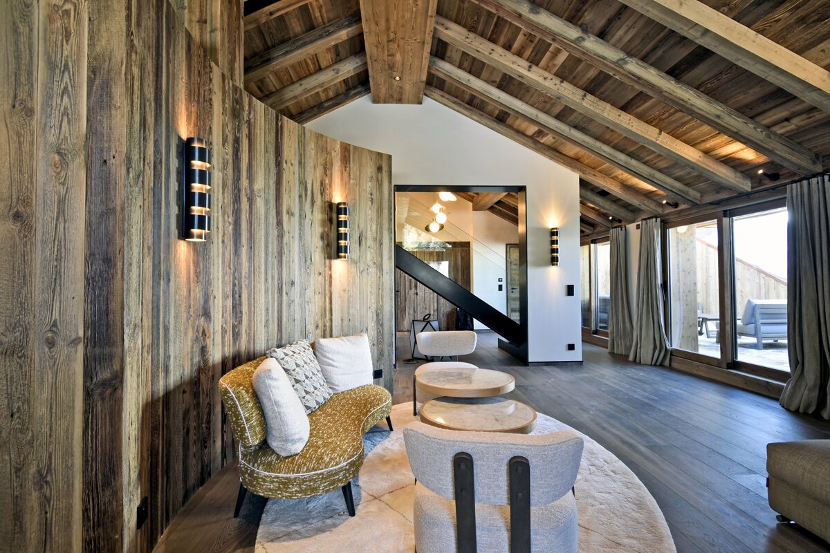 Chic chalet interior with rustic wood walls & vaulted ceiling, modern furnishings, warm lighting, and large windows revealing an outdoor area.