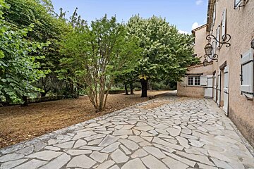 A stone patio leads past a beige building with white shutters and lanterns, toward a vibrant green garden with trees and dry, brown ground.