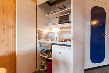 A compact kitchenette in an open white cupboard, featuring a sink, stove, microwave, and mini-fridge. Wood paneling on the left and a blue board on the right.