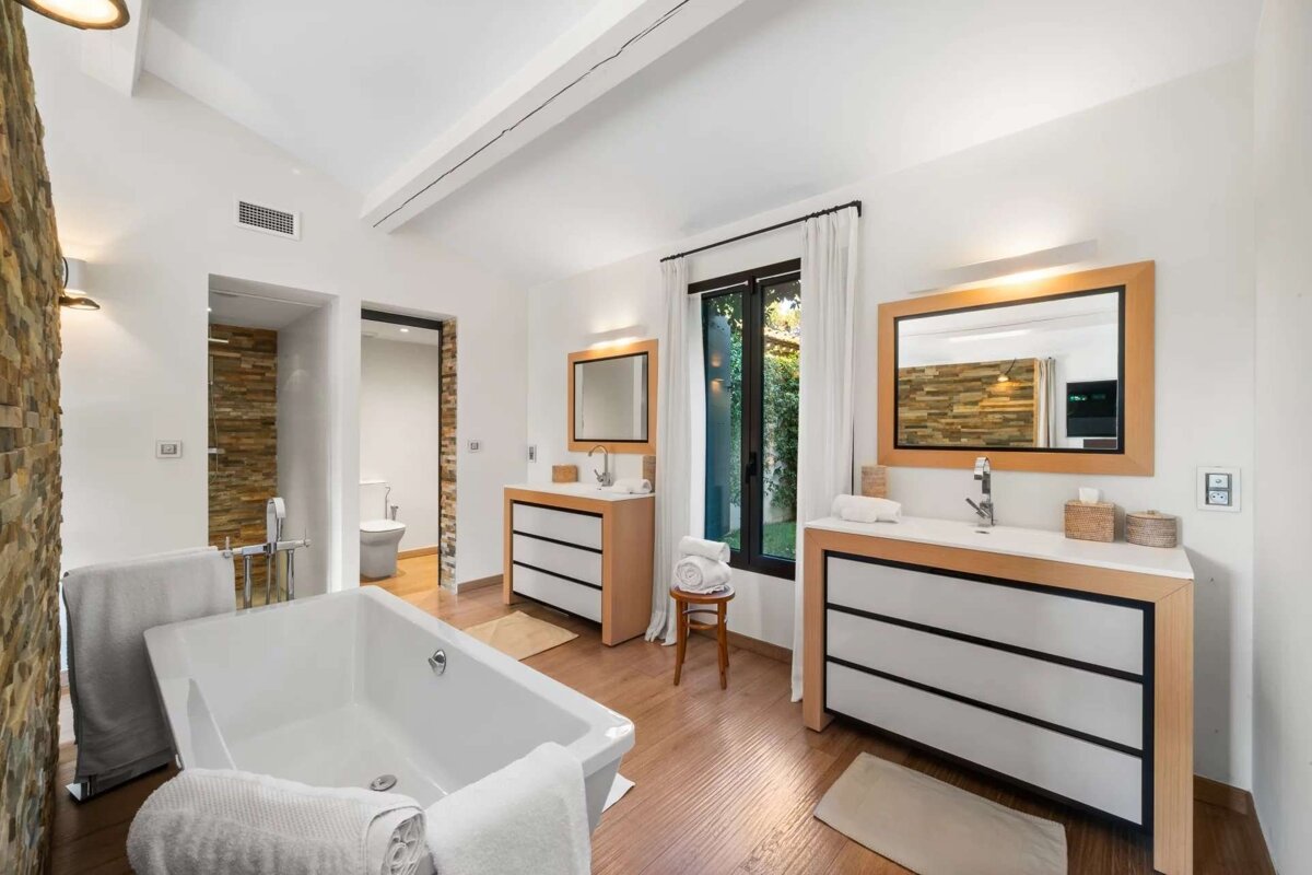 Bright, luxurious bathroom with a white tub, twin vanities, wooden floors, stone walls, and a large window with white curtains.