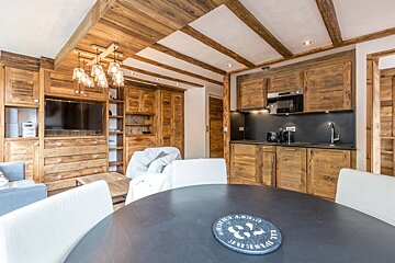 A warm, rustic chalet interior featuring extensive natural wood paneling, ceiling beams, an open-plan kitchen, dining area, and living room.