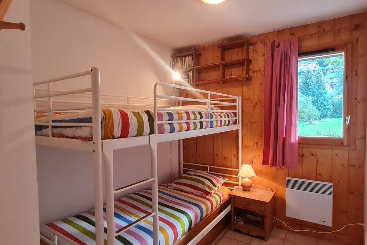 A cozy room features white bunk beds with colorful striped bedding, wood-paneled walls, a window with greenery outside, and a bedside lamp.