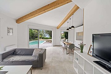 Bright, modern open-plan living/dining/kitchen with wood beams, grey sofa, and sliding doors leading to a pool and garden.