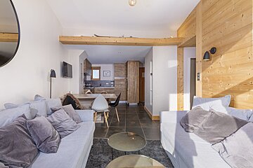 Modern chalet living room with light gray sofas, dark tiled floor, wooden walls & beams. Open kitchen/dining area in background, skis on beam.