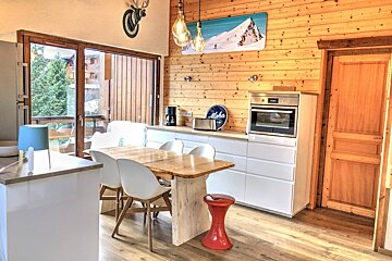 A modern kitchen/dining room with wood-paneled walls, white cabinets, a wooden table, and a balcony view. Features skiing art and a deer head.