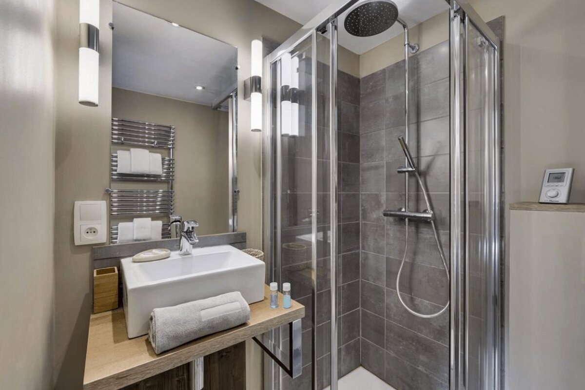 A modern bathroom featuring a white rectangular sink on a wooden counter and a glass-enclosed shower with grey tiling.
