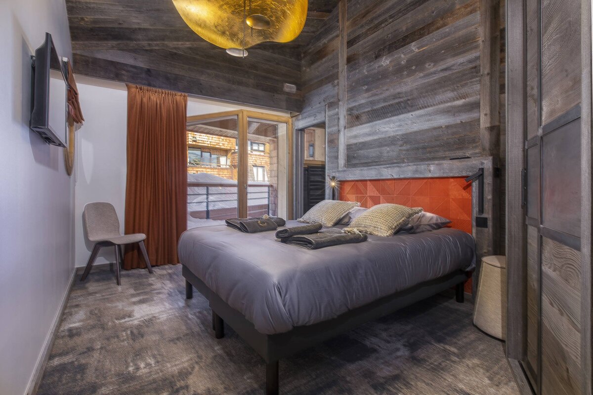 Rustic chalet bedroom featuring weathered wood walls, a grey double bed with an orange headboard, and a large gold ceiling light.