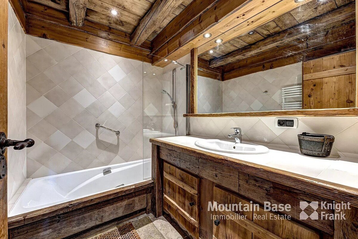 A rustic bathroom features exposed wooden beams, tiled walls, a bathtub with a shower, and a wooden vanity with a large mirror.