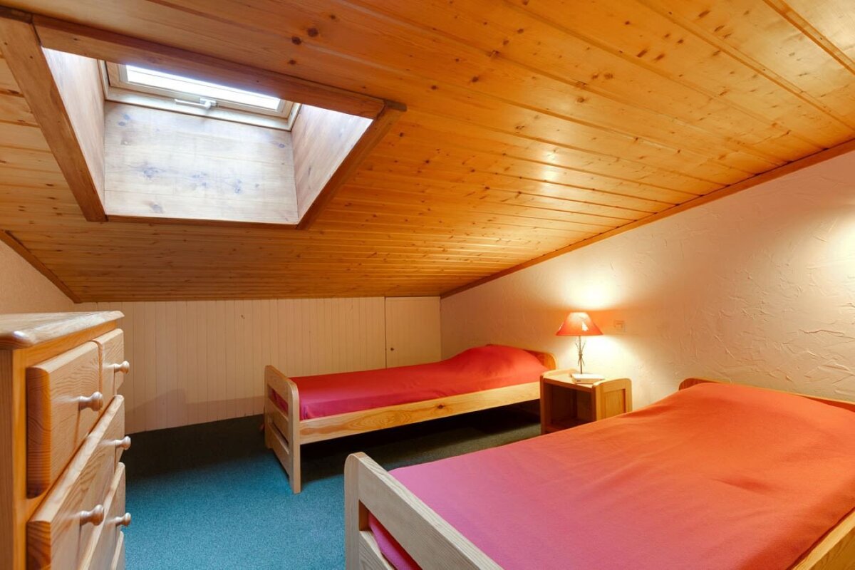 A cozy attic bedroom featuring a wood-paneled ceiling, a skylight, and two single beds with reddish bedding on a green carpet. A wooden dresser and lamp are visible.
