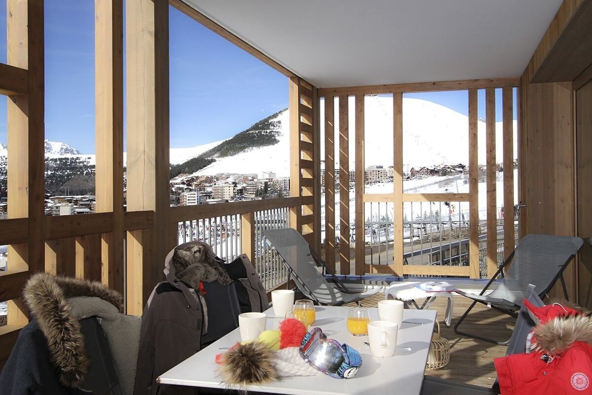 A cozy wooden balcony offers a sunny view of a snowy mountain ski resort. A table with drinks, ski goggles, hats, and winter jackets suggests a warm break.