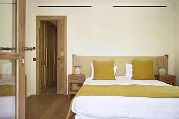 A bright bedroom with a large bed featuring a yellow headboard, pillows, and throw. Wooden floors, nightstands with lamps, and a wooden door create a warm, neutral ambiance.