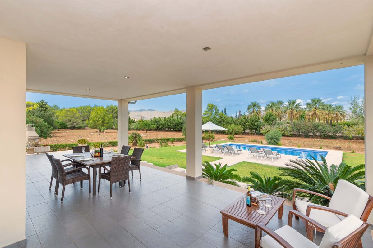 A spacious covered patio with dining and lounge areas overlooks a sunny garden with a swimming pool, lush greenery, and palm trees under a blue sky.