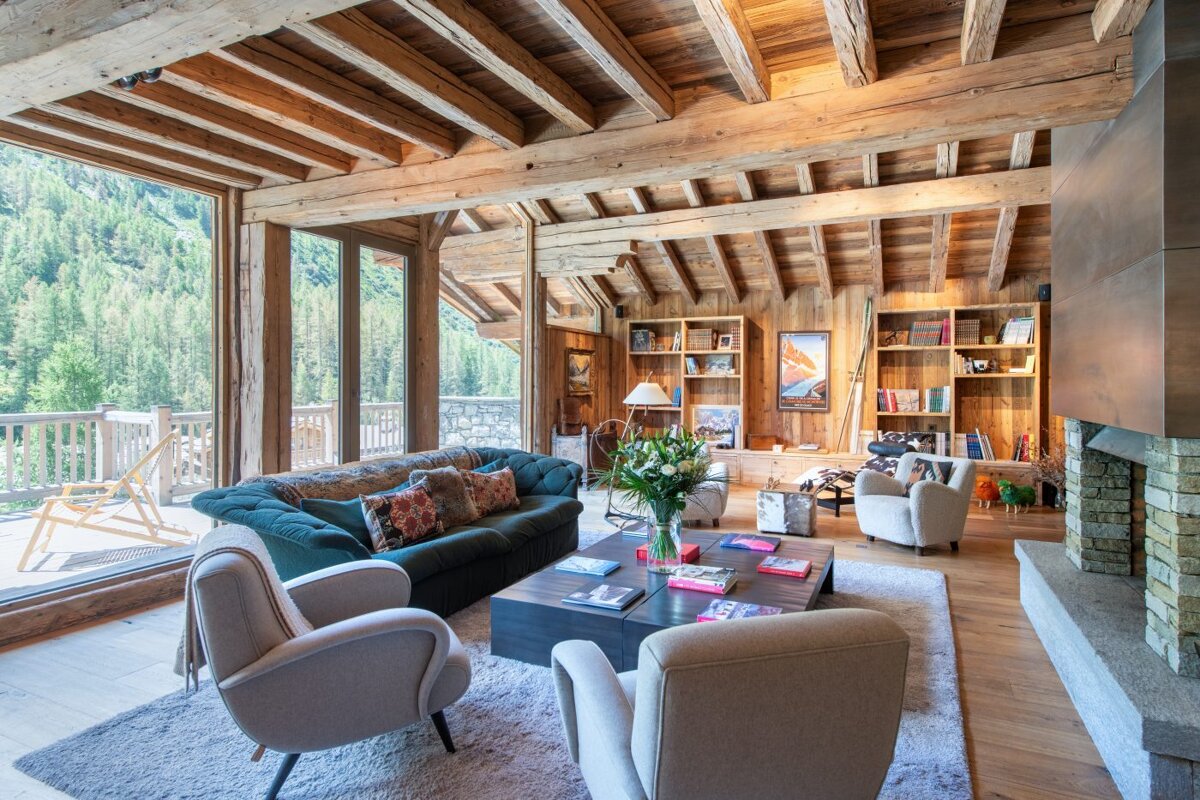 A cozy, rustic wooden chalet living room with a large sofa, armchairs, bookshelves, and a stone fireplace, offering expansive views of a lush forest.