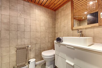 A compact bathroom featuring beige tiled walls, a wooden slat ceiling, modern white vanity with a rectangular sink, toilet, and radiator.