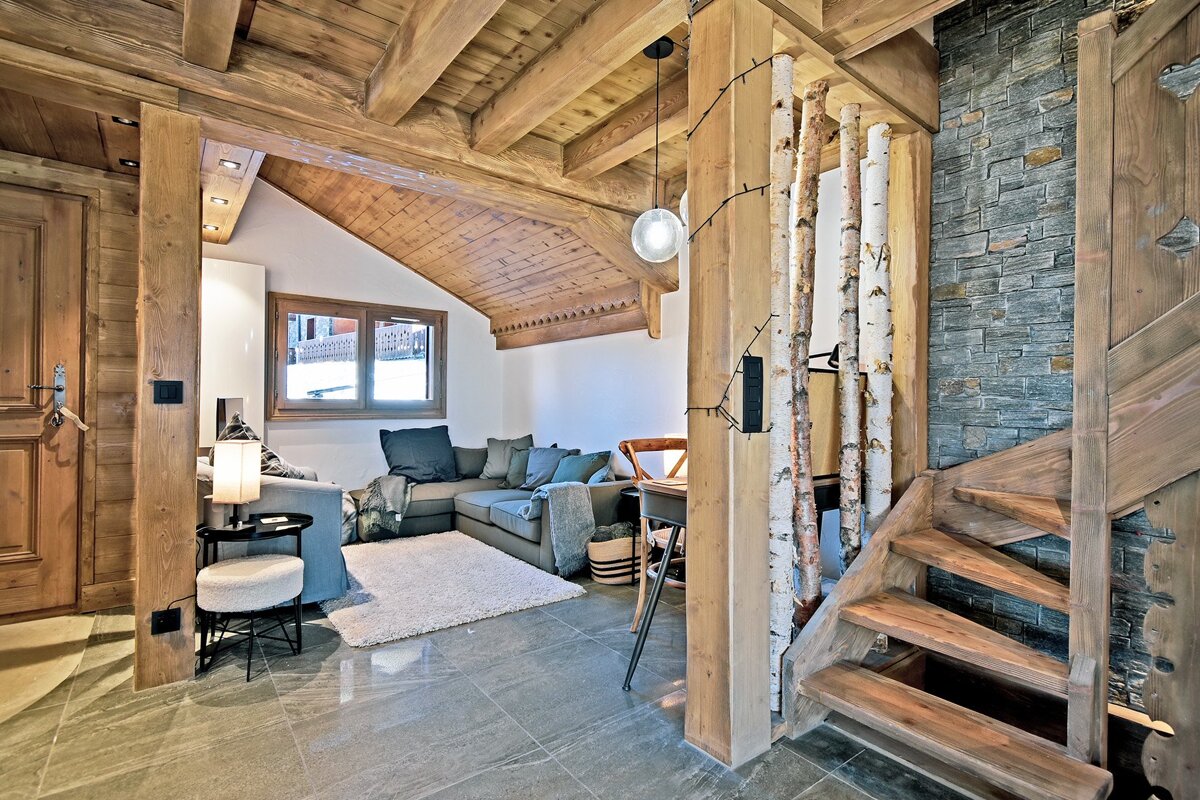 Cozy alpine chalet interior featuring wooden beams, a staircase, decorative birch logs, and a living area with a grey sectional sofa.