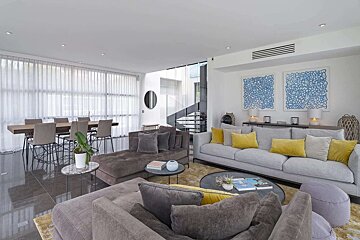 Bright, modern open-plan living/dining room with gray sofas, yellow pillows, a large dining table, and a striking spiral staircase. Blue abstract art.